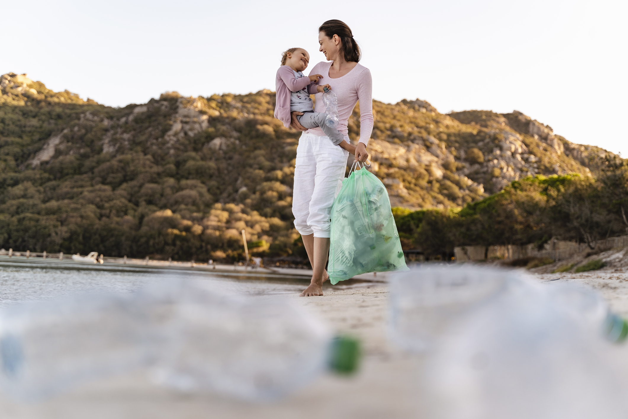 Woman on the beach carrying little daughter on her arm collecting empty plastic bottles